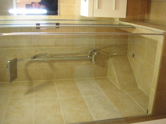 Kabina prysznicowa z beżowymi płytkami, widoczna armatura i szklane drzwi, odbicie osoby w lustrze w górnym rogu.