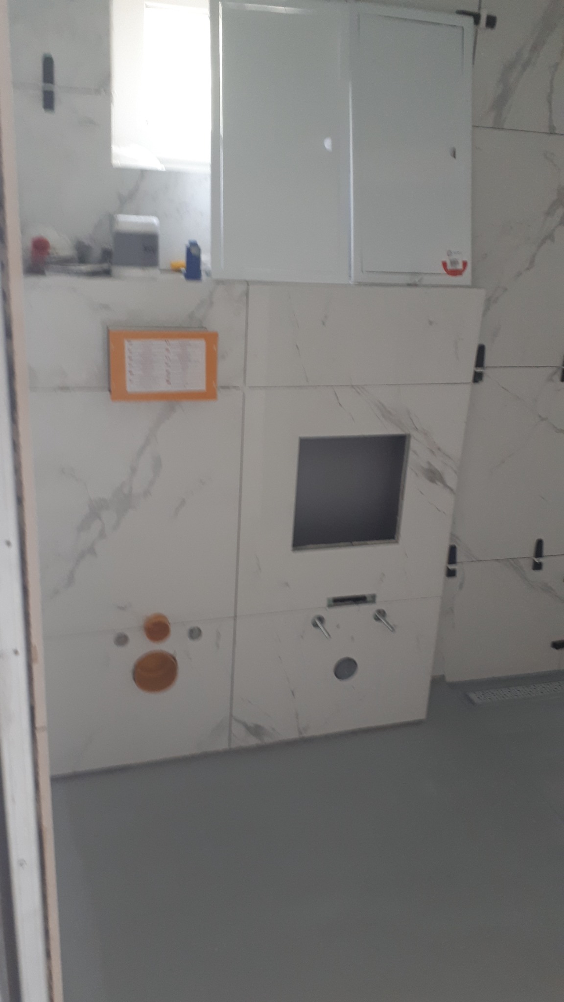 Świeżo wykafelkowana ściana w łazience z widocznymi przyłączami hydraulicznymi, otworem rewizyjnym i szafką nad stelażem podtynkowym WC, płytki imitujące marmur, szara podłoga.