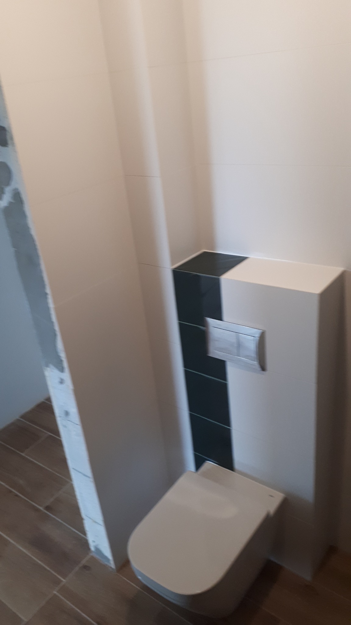 Podwieszana toaleta z zabudową w kolorze białym i czarnym, z chromowanym przyciskiem spłukującym, na tle ściany z jasnych płytek i drewnopodobnej podłogi; widoczny fragment ściany w stanie surowym.