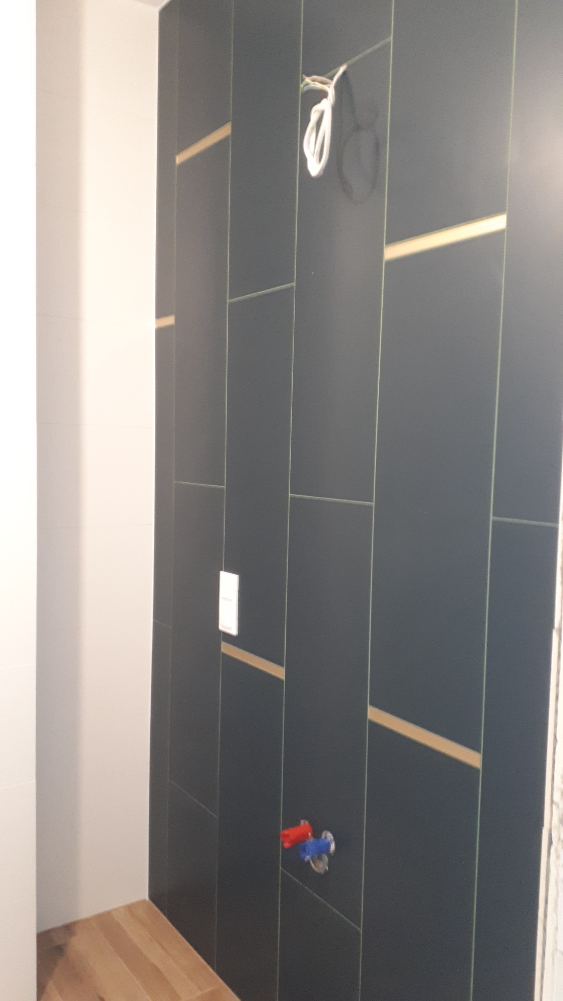 Ciemne, pionowo ułożone płytki w łazience z złotymi akcentami, widoczne wystające przewody i zawory wodne, fragment drewnopodobnej podłogi.