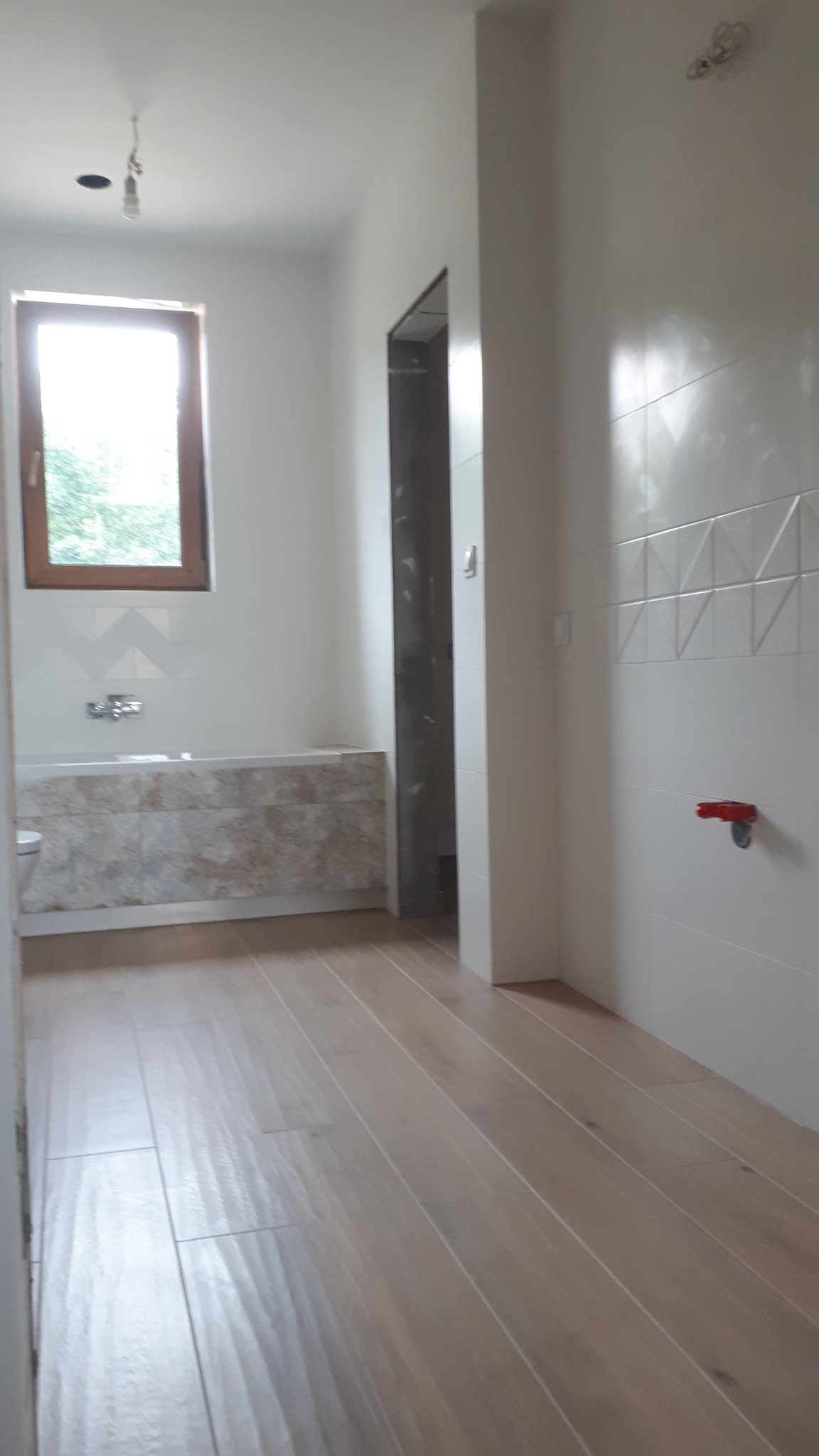 Wnętrze łazienki w trakcie prac wykończeniowych; widoczne drewnopodobne panele na podłodze, obudowana wanna z mozaiką oraz białe płytki z dekoracyjnym pasem na ścianach, brakujące elementy...