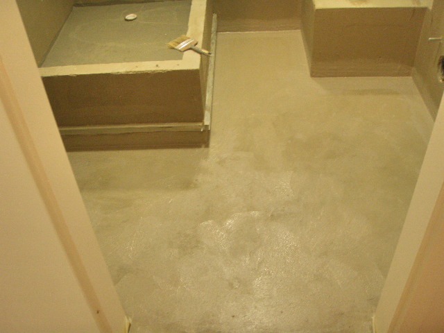 Niedokończona łazienka z nałożoną warstwą hydroizolacji na podłodze i w brodziku, widoczny pędzel malarski na krawędzi brodzika.