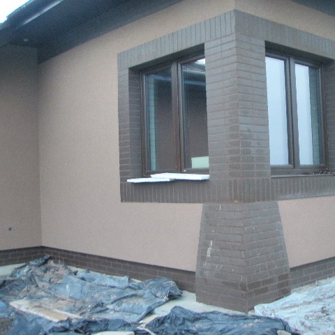 Elewacja domu jednorodzinnego z oknem obramowanym cegłą, widoczne zabezpieczenia folią malarską na ziemi.