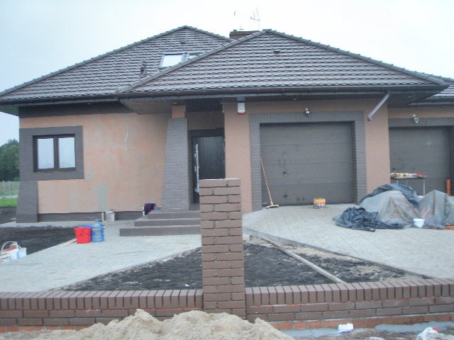 Dom jednorodzinny w trakcie prac wykończeniowych elewacji, widoczne świeżo położone tynki, częściowo wykończona ścieżka z kostki brukowej i murki z cegły klinkierowej w ogrodzie.