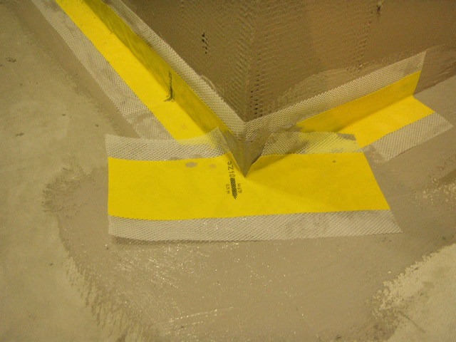 Hydroizolacja narożnika w łazience z użyciem żółtej taśmy uszczelniającej z białym brzegiem z siatki, na wilgotnej podłodze przed położeniem płytek.