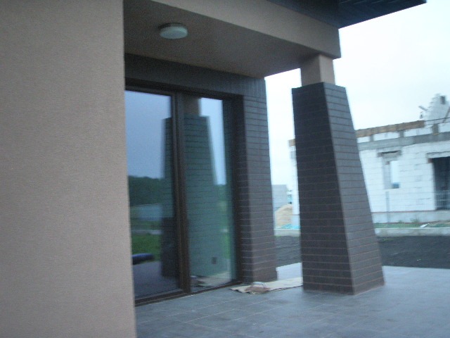 Wykończony fragment elewacji domu z brązowymi, ceglanymi filarami, przesuwnymi drzwiami tarasowymi i widoczną konstrukcją innego budynku w tle.