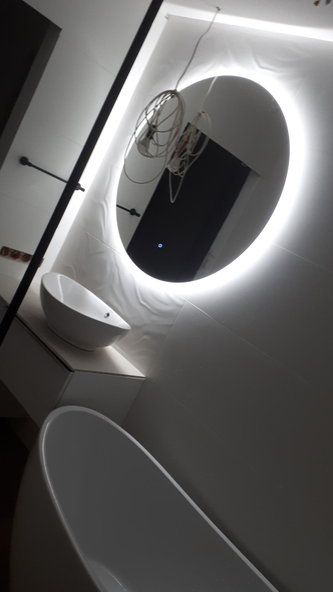 Nowoczesna łazienka w trakcie wykańczania: owalne lustro z podświetleniem LED, wisząca umywalka i fragment wanny, widoczne kable instalacji elektrycznej.