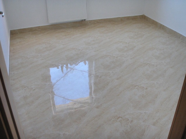 Jasne, polerowane płytki podłogowe z beżowym marmurkowym wzorem, odbijające światło okna, świeżo ułożone w pustym pomieszczeniu z białymi ścianami i grzejnikiem.