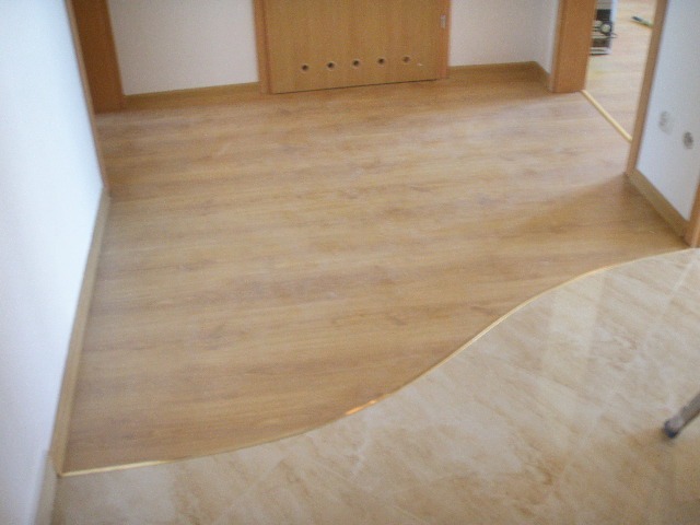 Jasne panele podłogowe imitujące drewno, połączone z jasnymi płytkami ceramicznymi za pomocą ozdobnej, złotej listwy, widoczne drzwi w tle.