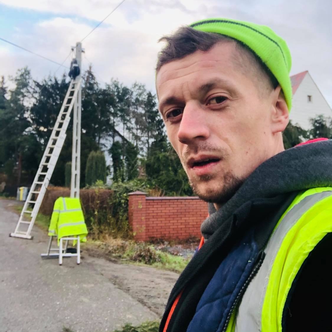 Autoportret elektryka w neonowej czapce i kamizelce odblaskowej, w tle słup elektryczny z pracownikiem na drabinie oraz kurtka na krześle