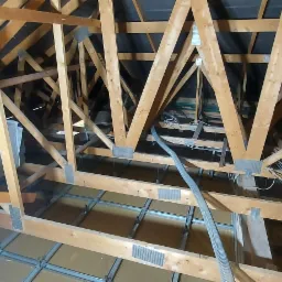 Drewniana konstrukcja dachu poddasza z widoczną warstwą izolacyjną i przewodami elektrycznymi, częściowo oświetlona naturalnym światłem.