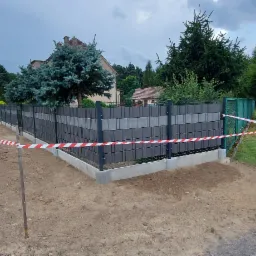 Nowoczesne ogrodzenie z paneli betonowych w odcieniach szarości, z widocznymi słupkami i podmurówką, zabezpieczone taśmą ostrzegawczą na tle zieleni i domu.