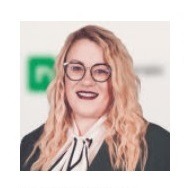 Portret kobiety w okularach, z blond włosami i w ciemnej marynarce z białą bluzką, uśmiechającej się na tle zielonego logo biura nieruchomości.