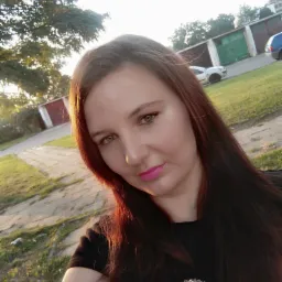 Portret kobiety z długimi, ciemnymi włosami i różową szminką na tle garaży i zieleni w słoneczny dzień.