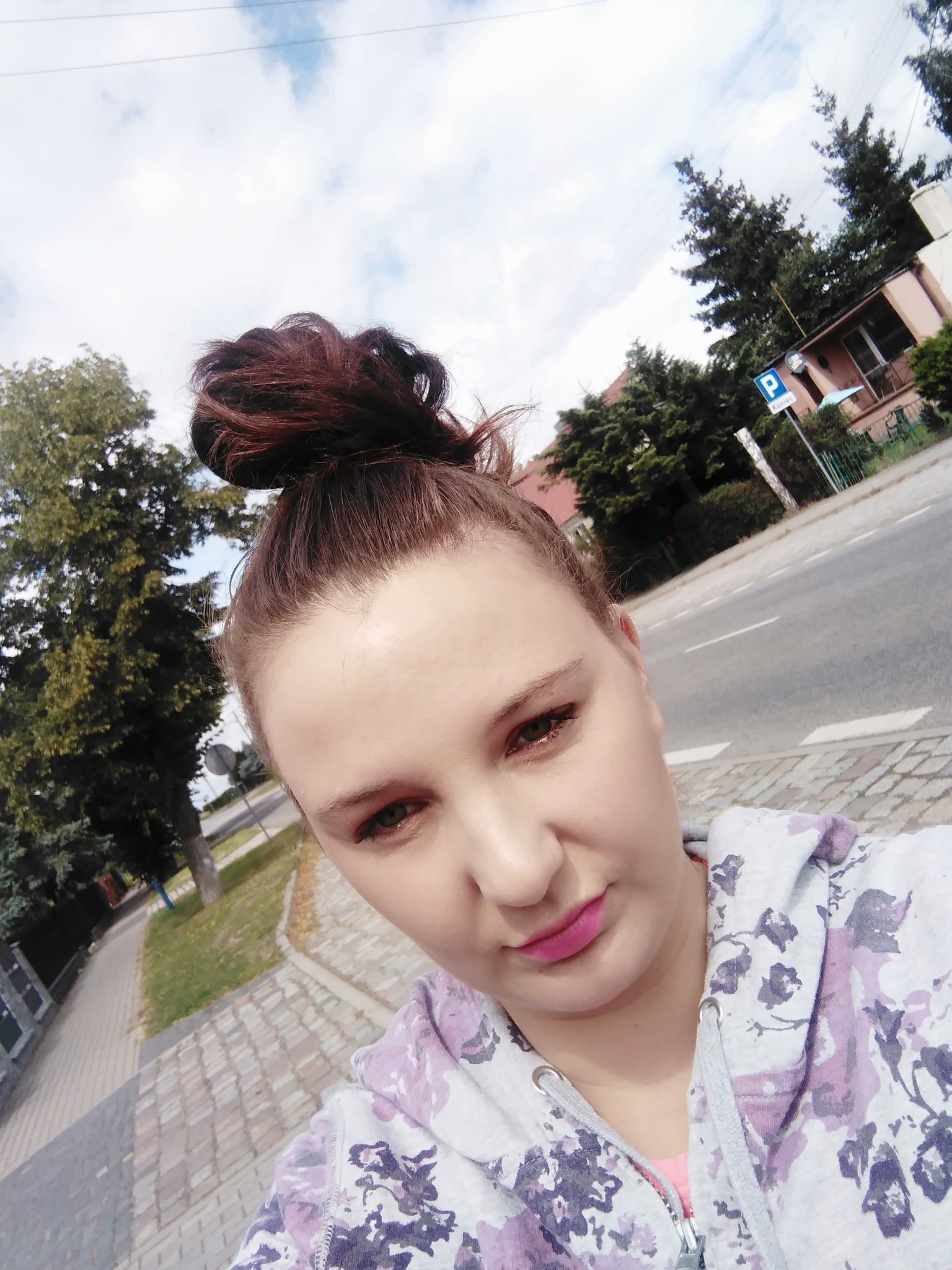 Portret kobiety z brązowymi włosami upiętymi w kok, ubrana w bluzę z kapturem w kwiatowy wzór, stojącej na chodniku z kostki brukowej na tle ulicy i zieleni.