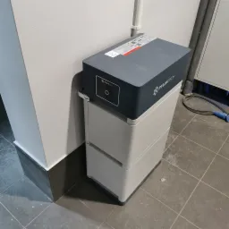 Szara bateria magazynu energii Pylontech, widok z boku, na szarej podłodze z płytek, obok białej ściany z ciemnoszarą listwą przypodłogową.