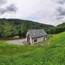 Nowoczesny, parterowy dom z szarym dachem i drewnianymi elementami wykończeniowymi, zlokalizowany na zielonym wzgórzu w otoczeniu lasu, z białym vanem zaparkowanym w pobliżu drewnianego ogrodzenia.