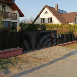 Nowoczesna, pozioma, brązowa brama wjazdowa z metalowymi lamelami, wkomponowana w czarne, metalowe ogrodzenie na niskiej, ceglanej podmurówce, na tle dwóch domów.