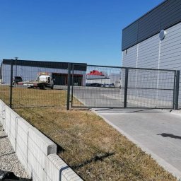 DS PŁOT - TRANS SYLWIA MARSZAŁKOWSKA - Szara brama przemysłowa z paneli siatkowych zamontowana przed budynkiem magazynowym, widoczny holownik na trawniku w tle.