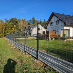 DS PŁOT - TRANS SYLWIA MARSZAŁKOWSKA - Długie, szare, betonowe podmurówki z fakturą kamienia podtrzymują panele z ciemnego drutu, tworząc ogrodzenie posesji z nowoczesnymi domami w tle, widoczne cień fotografa.