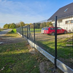 DS PŁOT - TRANS SYLWIA MARSZAŁKOWSKA - Czarny ogrodzenie panelowe z podmurówką betonową, widok z boku, w tle dom z czerwonym samochodem osobowym zaparkowanym obok.