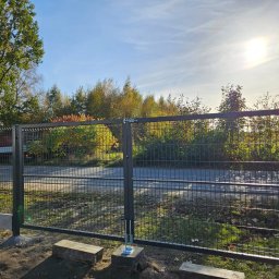 DS PŁOT - TRANS SYLWIA MARSZAŁKOWSKA - Nowa, ciemnoszara brama panelowa z widocznym mechanizmem otwierania, zamontowana na betonowej podmurówce, z zielenią w tle i fragmentem samochodu dostawczego po lewej stronie.