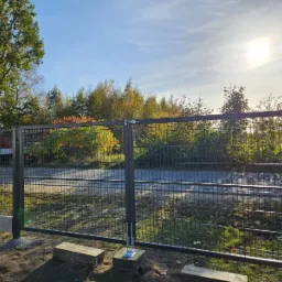 Nowa, ciemnoszara brama panelowa z widocznym mechanizmem otwierania, zamontowana na betonowej podmurówce, z zielenią w tle i fragmentem samochodu dostawczego po lewej stronie.