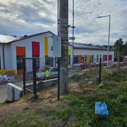 DS PŁOT - TRANS SYLWIA MARSZAŁKOWSKA - Nowoczesne ogrodzenie panelowe w kolorze antracytowym, z widocznym słupem energetycznym i fragmentem budynku z kolorowymi akcentami, teren w trakcie prac wykończeniowych.