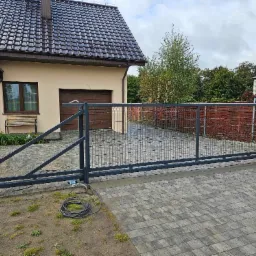 Automatyczna brama przesuwna z metalową siatką na posesji z kostki brukowej, widoczny silnik i zwinięty kabel zasilający obok, dom z brązowym dachem w tle.