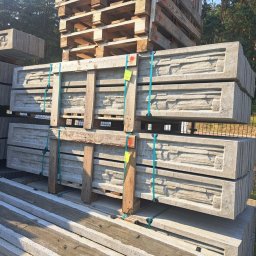 DS PŁOT - TRANS SYLWIA MARSZAŁKOWSKA - Betonowe elementy ogrodzeniowe z dekoracyjnym wzorem ułożone na paletach i zabezpieczone taśmami, widoczne w składzie budowlanym na tle drzew.