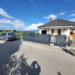 Nowoczesna posesja z przesuwną bramą w kolorze grafitowym i ogrodzeniem z poziomych profili, widok z ulicy, na pierwszym planie cień osoby robiącej zdjęcie.