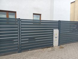 Nowoczesne, poziome przęsła w kolorze grafitowym z białymi akcentami, tworzące ogrodzenie posesji. Widoczna skrzynka elektryczna wkomponowana w ogrodzenie. Tło stanowi biały budynek z brązowymi...