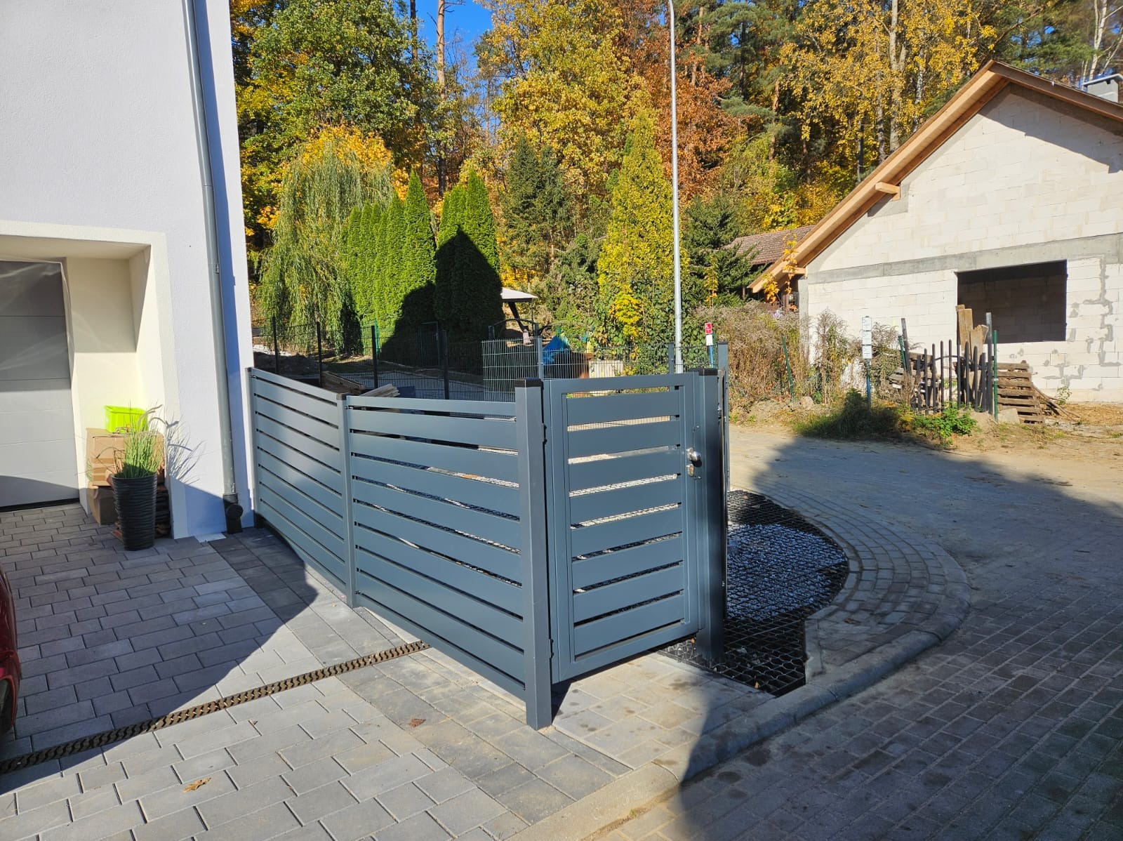 Nowoczesne, szare ogrodzenie z poziomych paneli metalowych, oddzielające posesję od ulicy z kostki brukowej, w tle budynek w stanie surowym i drzewa z jesiennymi liśćmi.