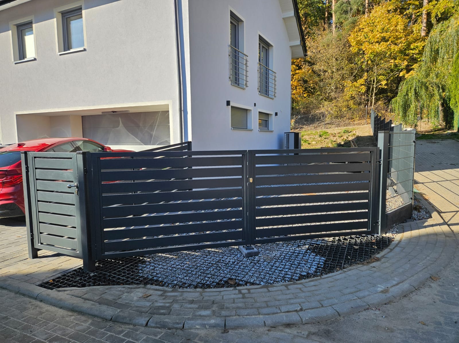 Nowoczesna, grafitowa brama wjazdowa z poziomymi profilami, zamontowana na posesji z jasnym domem i widocznym fragmentem czerwonego samochodu w tle, na podłożu z siatki i drobnego kamienia.