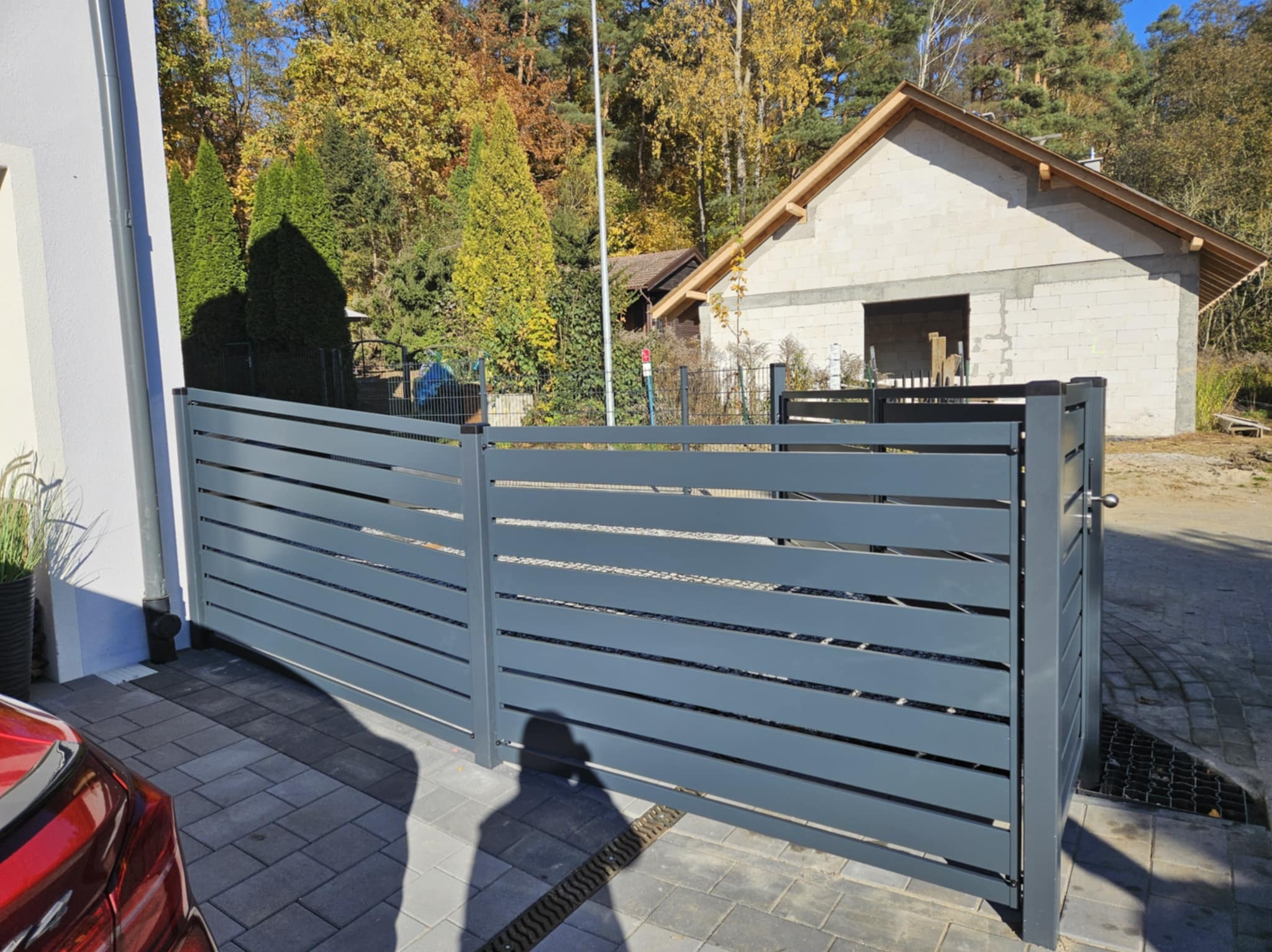 Nowoczesna, przesuwna brama w kolorze grafitowym z poziomymi panelami, zamontowana na posesji z kostki brukowej, w tle budynek w stanie surowym otwartym i drzewa.