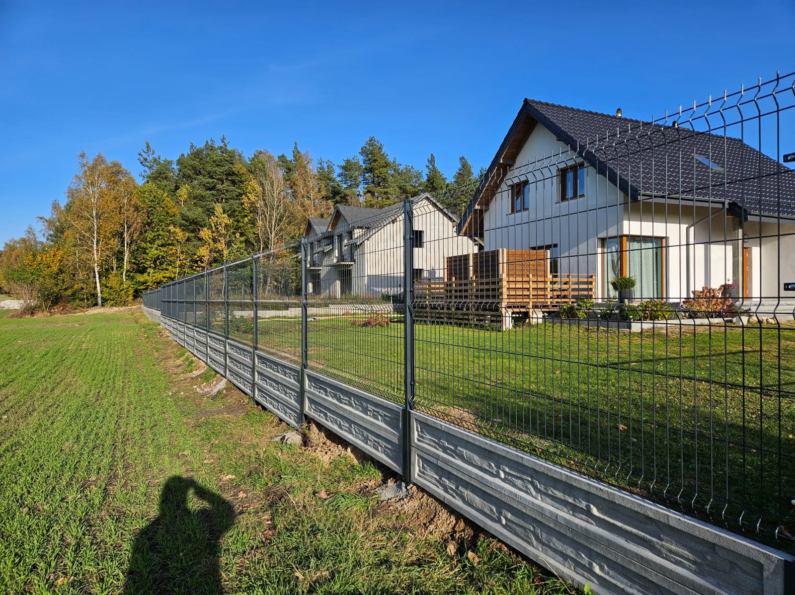 Długie, szare, betonowe podmurówki z fakturą kamienia podtrzymują panele z ciemnego drutu, tworząc ogrodzenie posesji z nowoczesnymi domami w tle, widoczne cień fotografa.