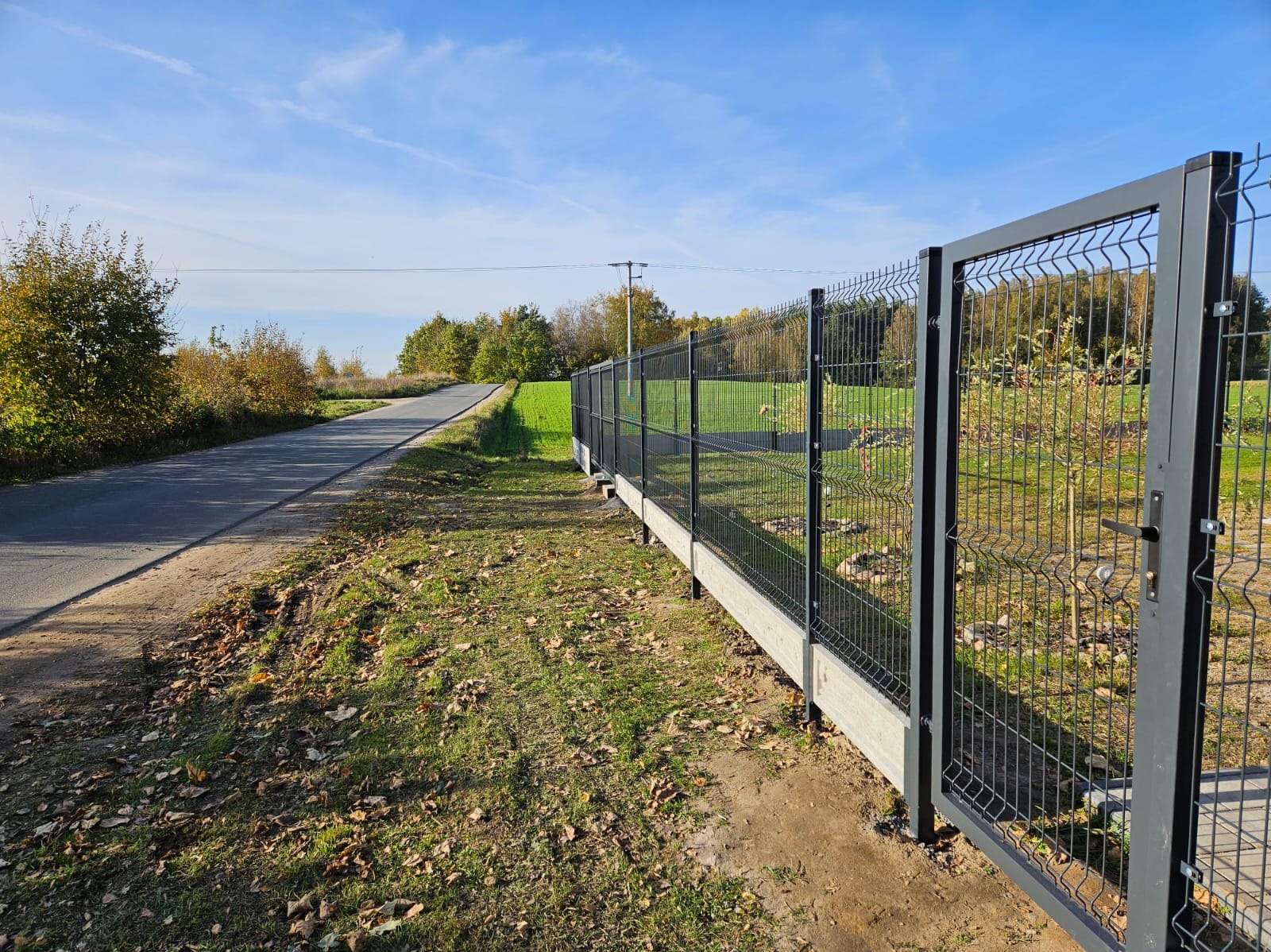 Nowoczesne ogrodzenie panelowe z bramą, osadzone na betonowej podmurówce, widoczne z perspektywy drogi wiejskiej w słoneczny dzień.