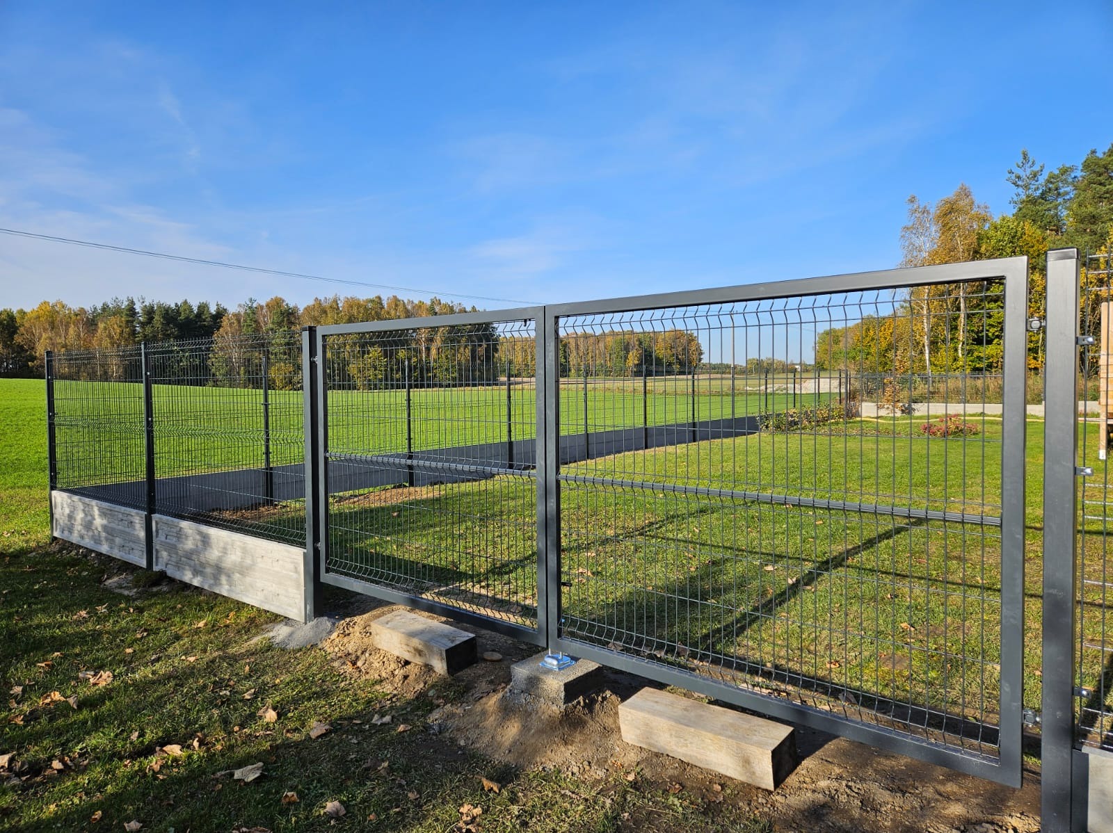 Nowoczesne, grafitowe ogrodzenie panelowe z bramą dwuskrzydłową, osadzone na betonowej podmurówce, widoczne na tle zielonego trawnika i drzew w jesiennej scenerii.