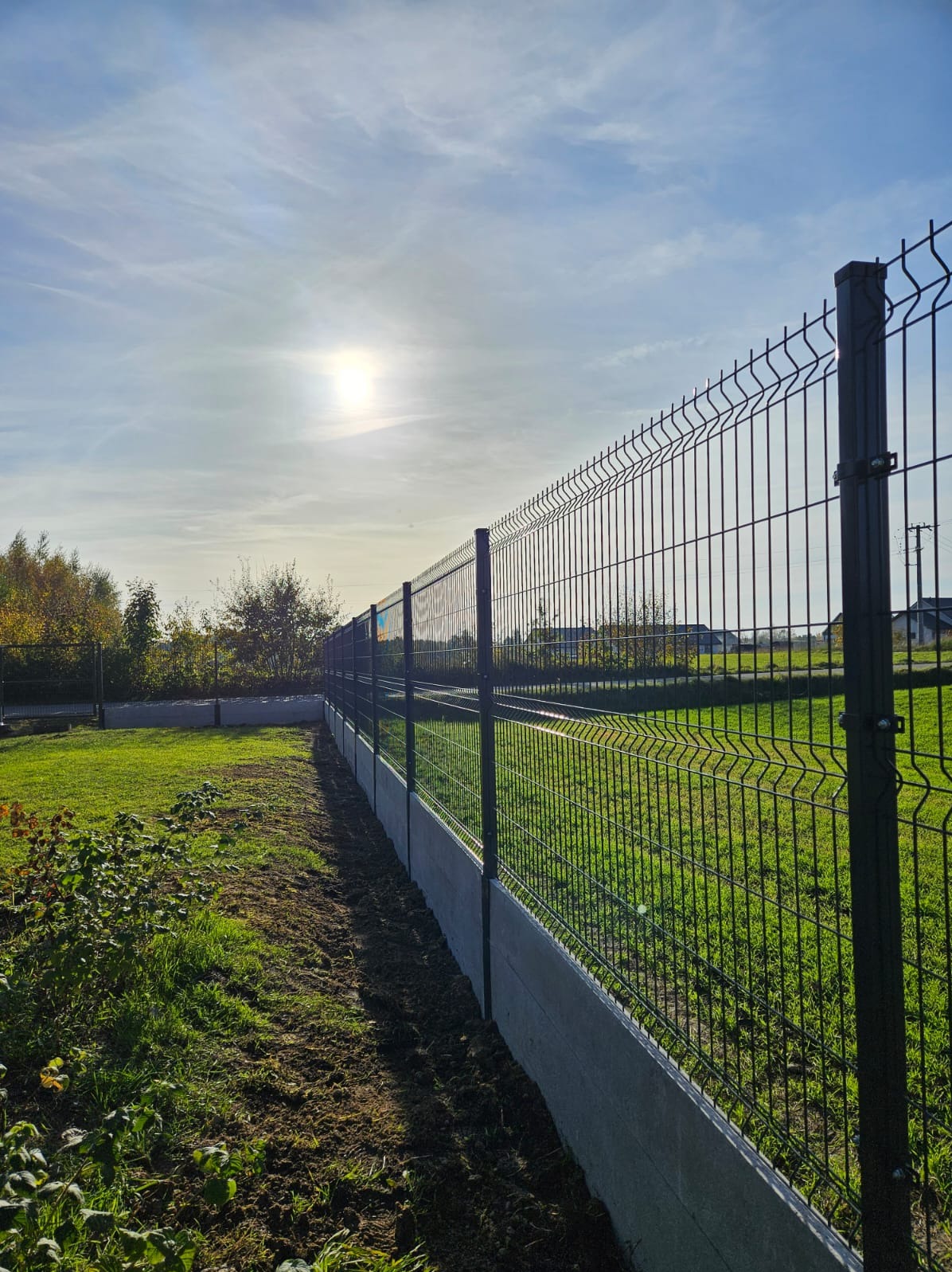 Długi, ciemny panel ogrodzeniowy na betonowej podmurówce, świeżo obsypany ziemią, z widokiem na trawnik i zabudowania w oddali, pod błękitnym, słonecznym niebem.