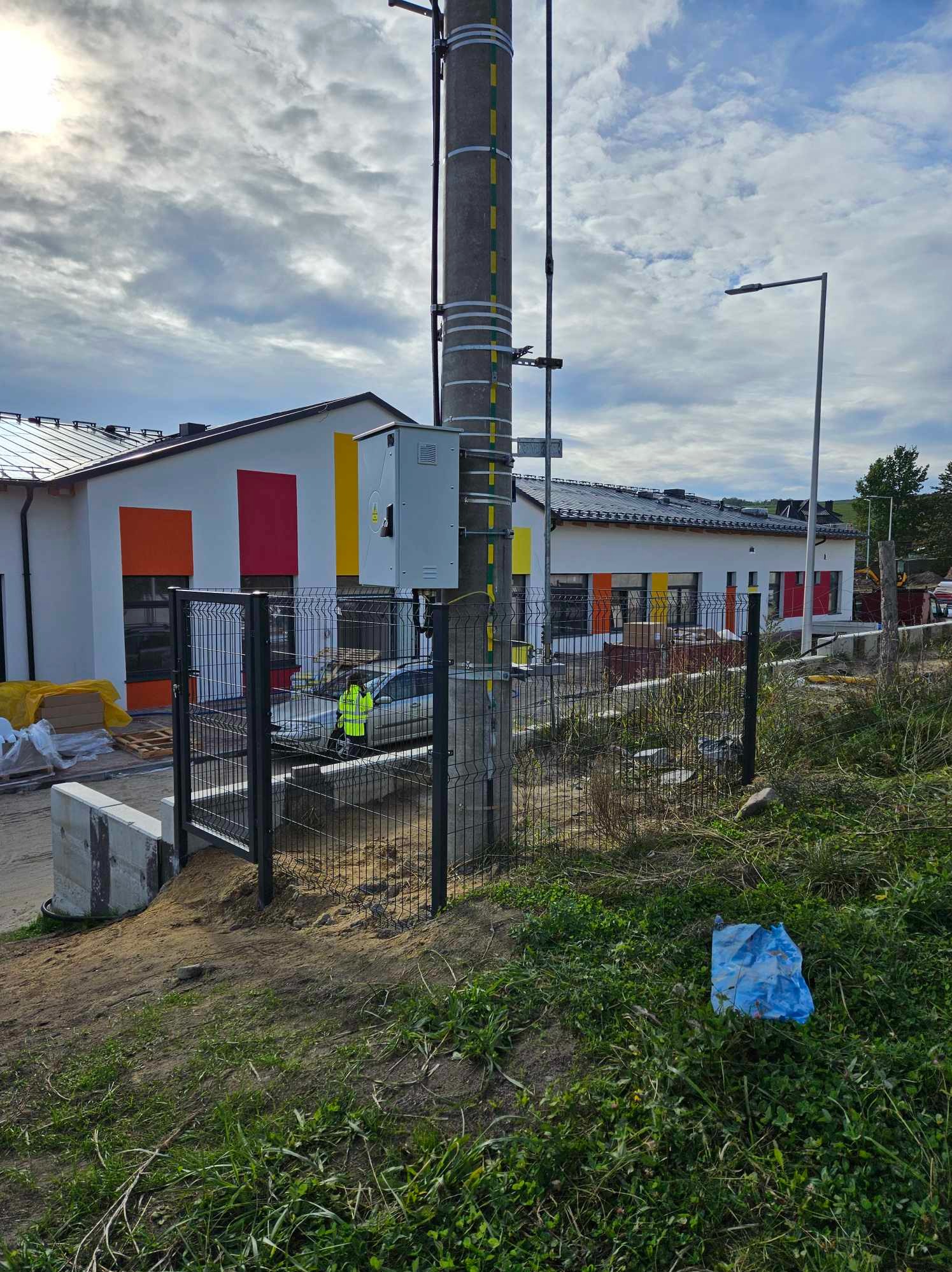Nowoczesne ogrodzenie panelowe w kolorze antracytowym, z widocznym słupem energetycznym i fragmentem budynku z kolorowymi akcentami, teren w trakcie prac wykończeniowych.