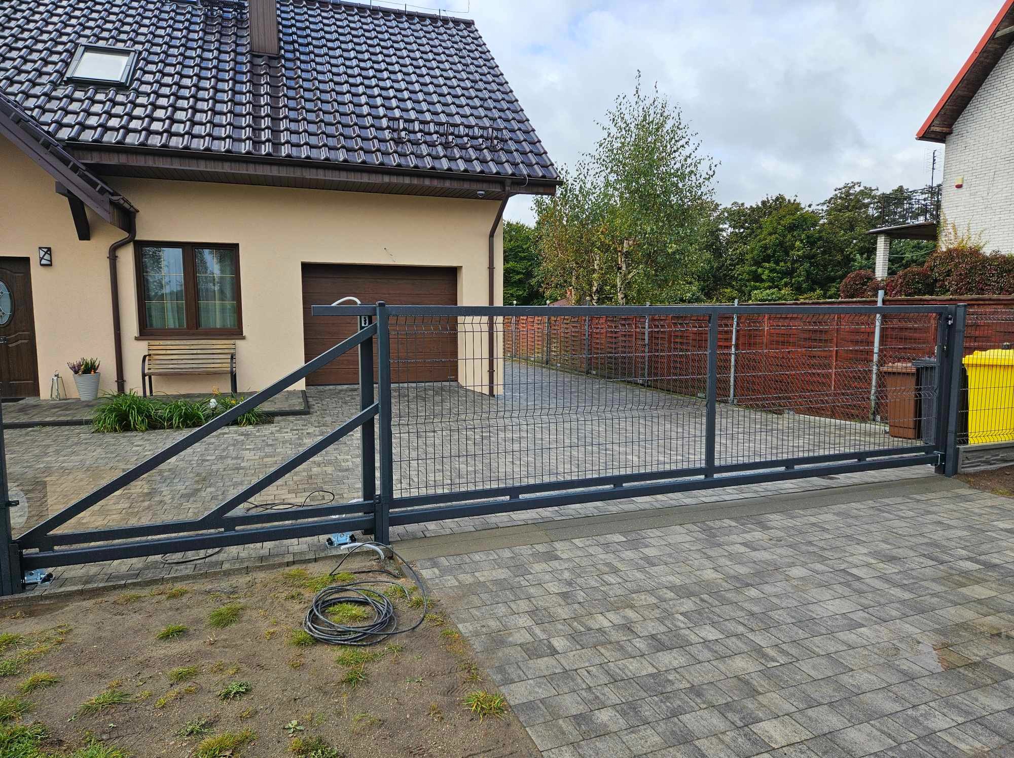 Automatyczna brama przesuwna z metalową siatką na posesji z kostki brukowej, widoczny silnik i zwinięty kabel zasilający obok, dom z brązowym dachem w tle.
