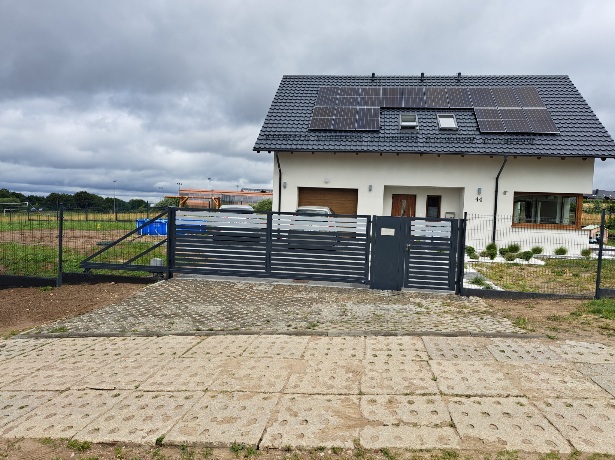 Nowoczesna posesja z szarą, przesuwną bramą i ogrodzeniem panelowym, widok z zewnątrz na dom z panelami słonecznymi na dachu w pochmurny dzień.