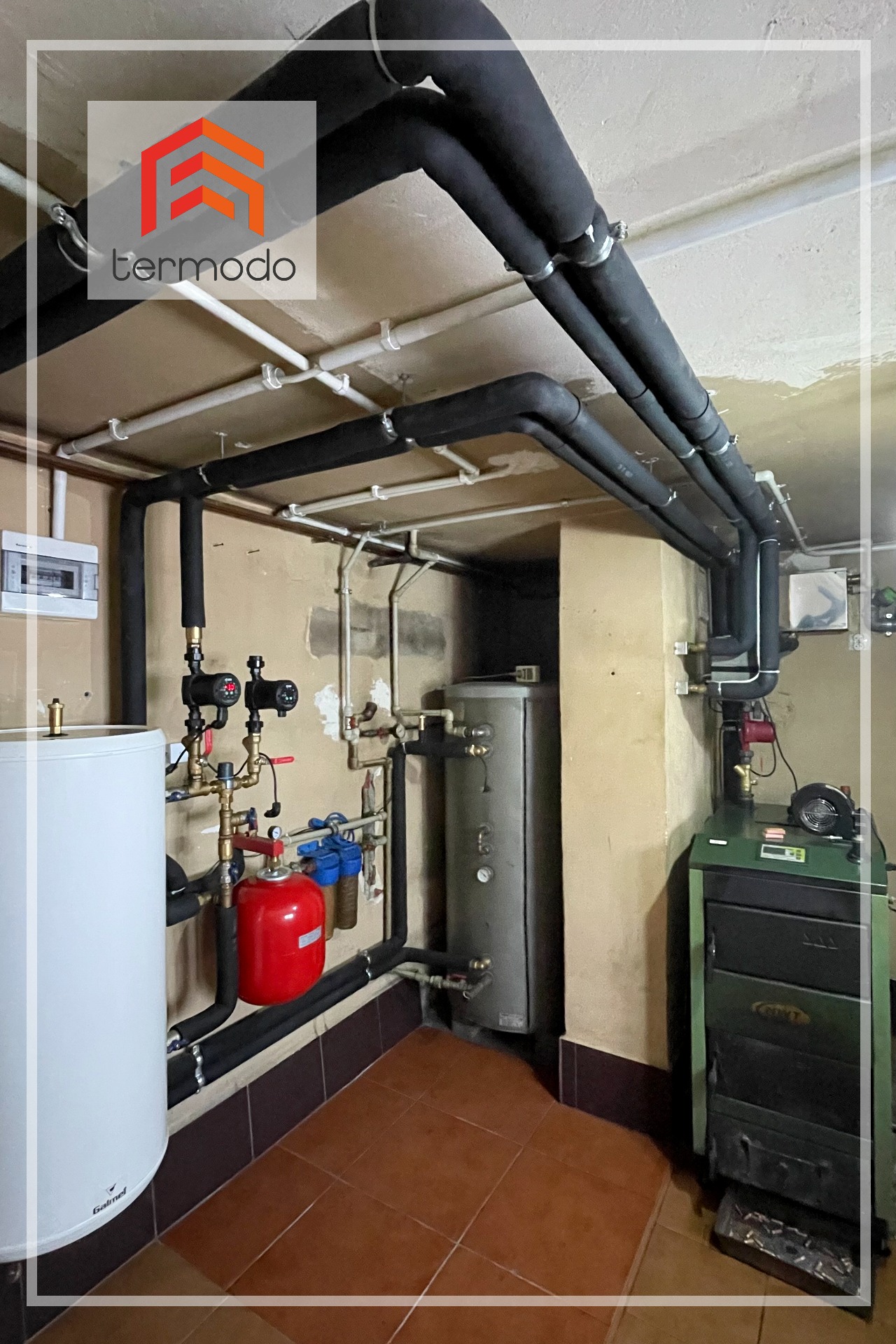 Hybrydowy system grzewczy z kotłem stałopalnym i instalacją rur w izolacji, widoczny zbiornik buforowy, naczynie wzbiorcze i zielony kocioł, na ścianie zamontowana skrzynka elektryczna