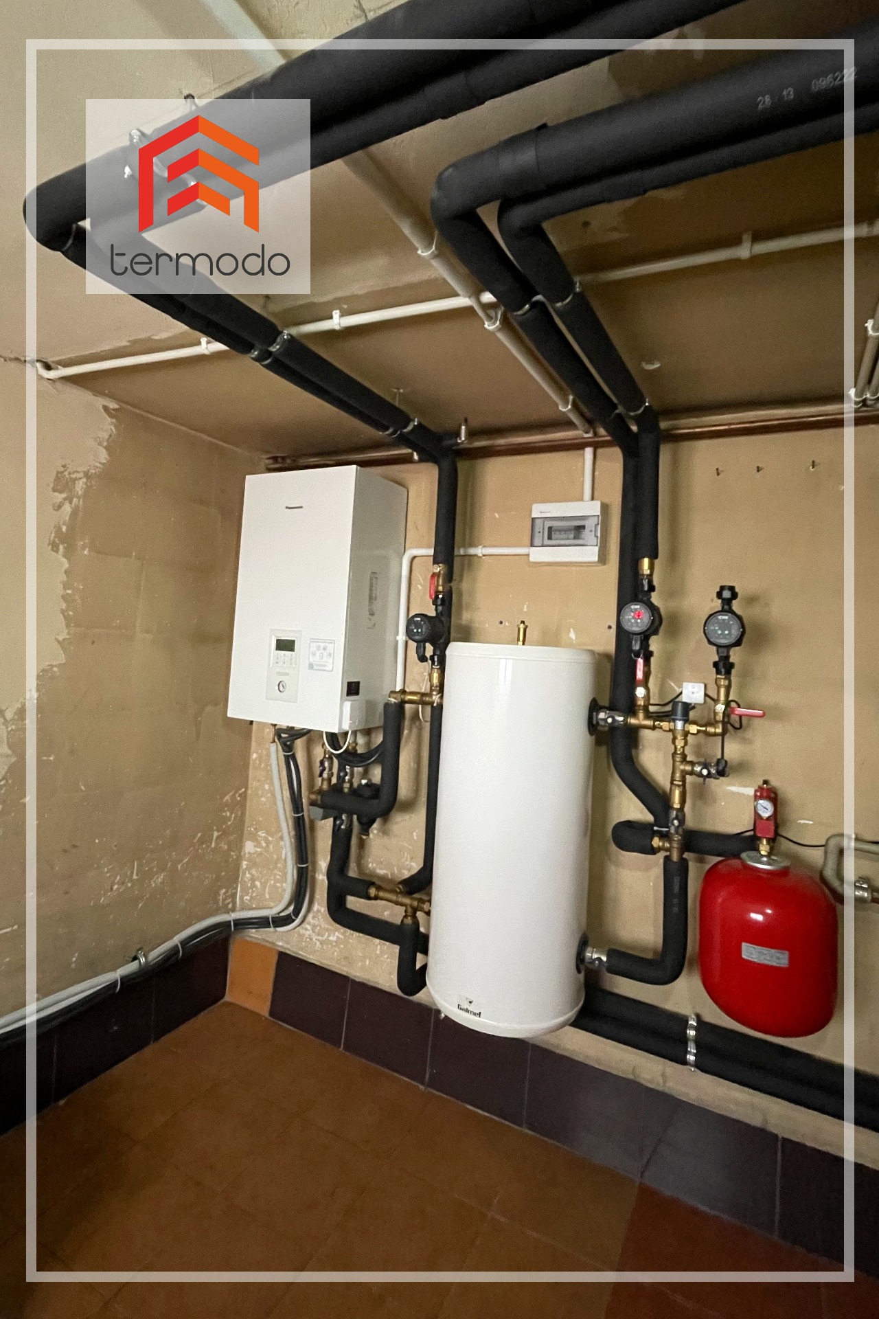 Hybrydowa instalacja grzewcza: Pompa ciepła Panasonic T-Cap SQC 9kW połączona z kotłem stałopalnym, widoczne czarne rury izolacyjne i czerwony zbiornik wyrównawczy w piwnicy.