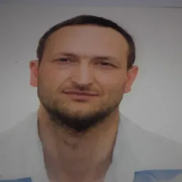 Portret mężczyzny z krótką, ciemną brodą i włosami, ubrany w jasną koszulę, na białym tle.