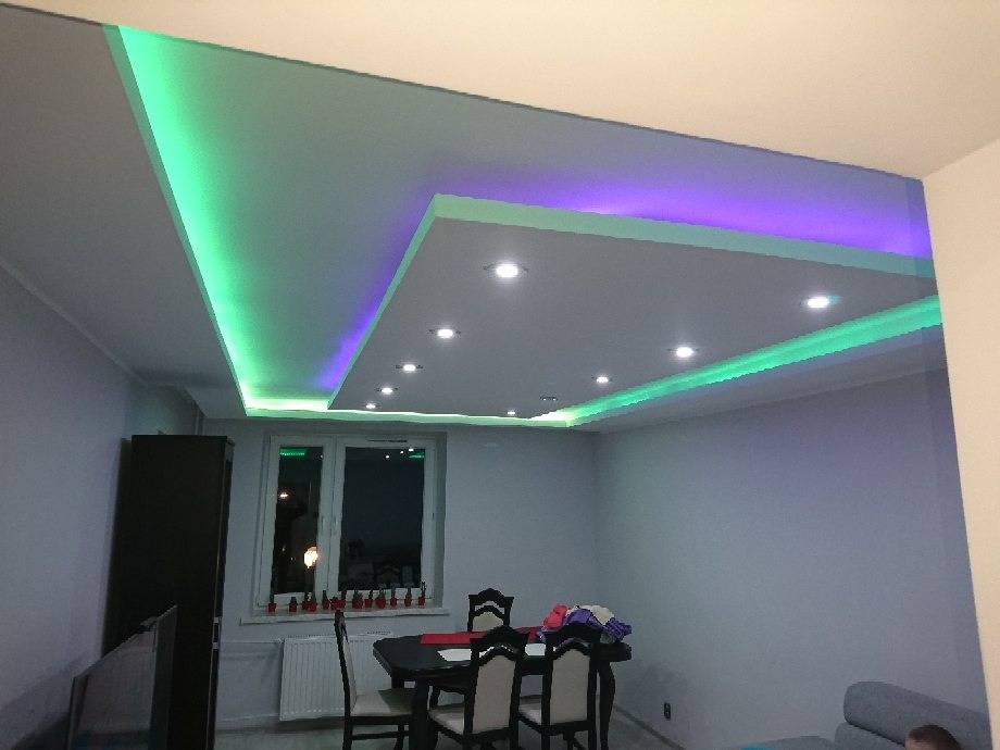 Nowoczesny salon z podwieszanym sufitem z oświetleniem LED w kolorach zielonym i fioletowym, widok na stół jadalny i okno.