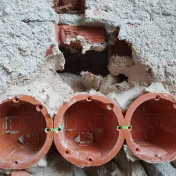 Trzy pomarańczowe puszki elektryczne zamontowane w surowej, otynkowanej ścianie z widocznymi fragmentami cegieł, wyrównane za pomocą lasera krzyżowego.