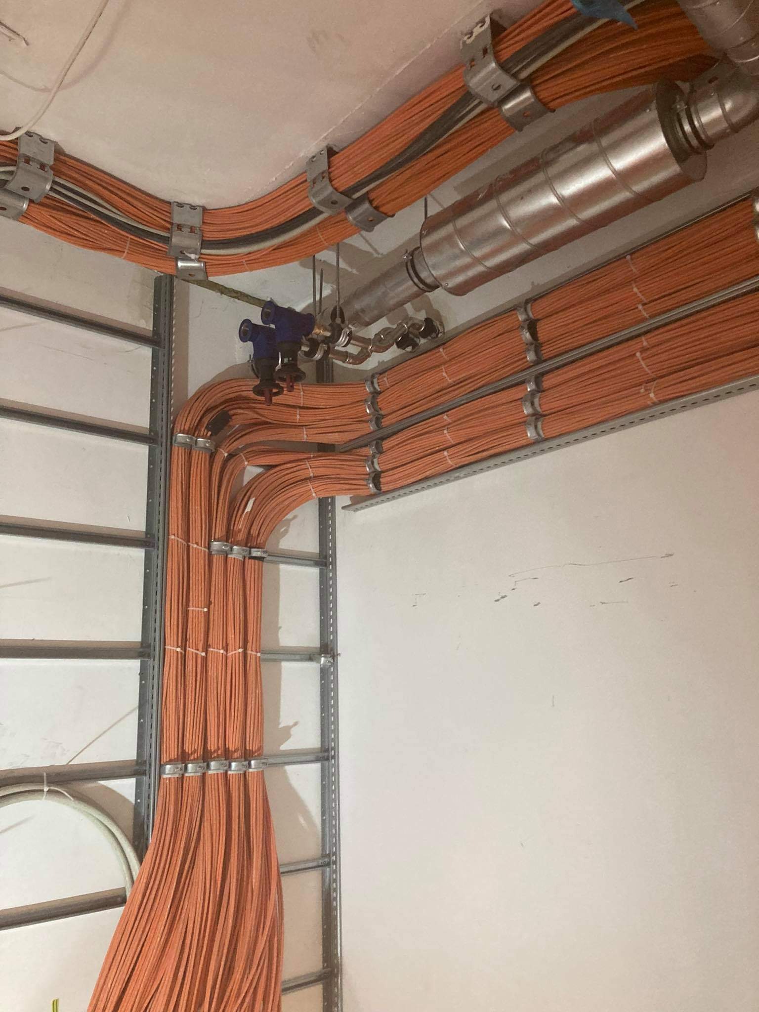 Pomarańczowe przewody elektryczne ułożone w korytach kablowych, zamocowane do ściany i sufitu, widoczne rury instalacji wodnej.