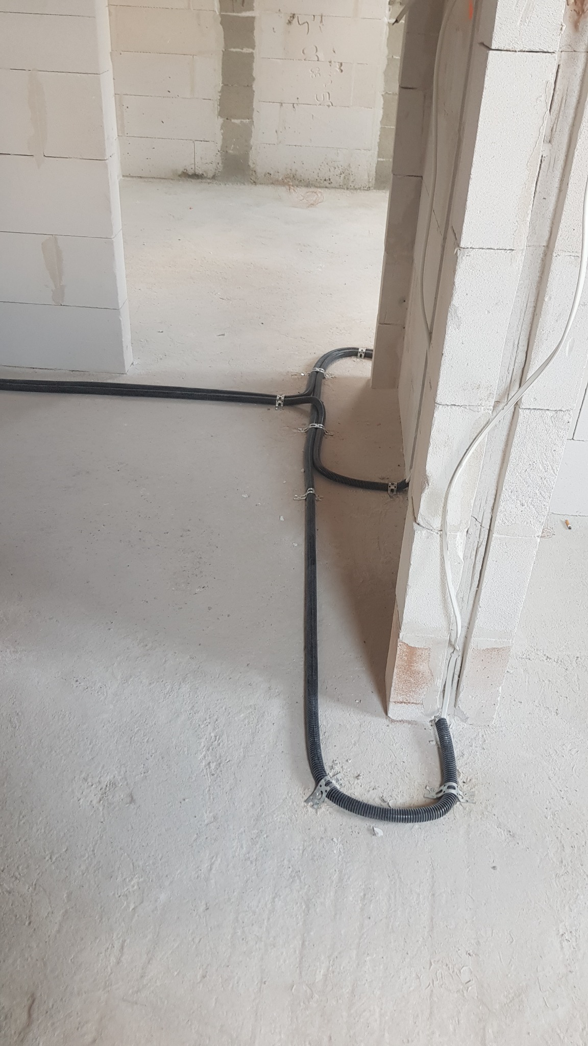 Czarna, karbowana rura kablowa przymocowana metalowymi uchwytami do surowej ściany z betonu komórkowego podczas prac remontowych.