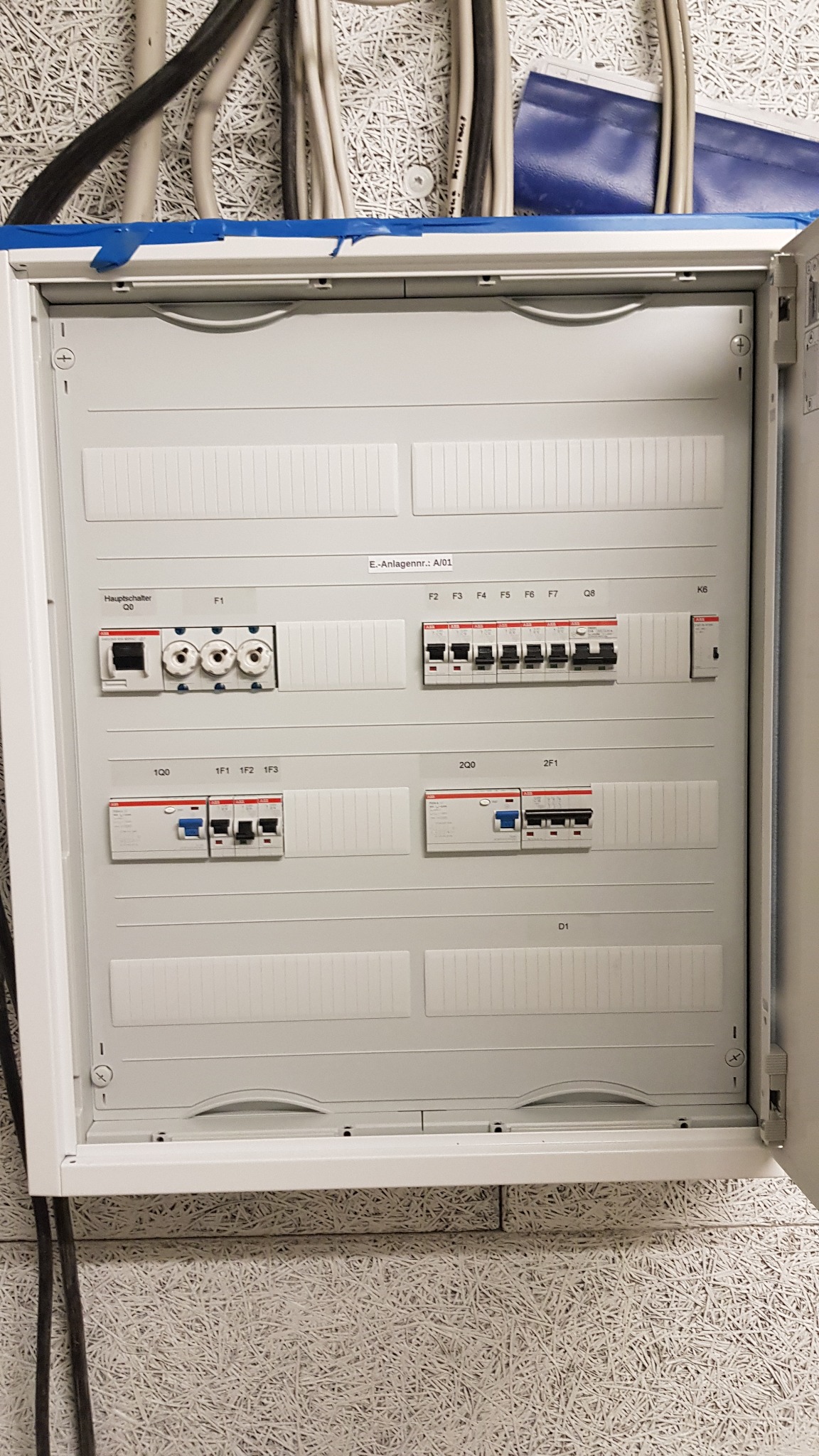 Otwarta szafa elektryczna z bezpiecznikami i okablowaniem, zamontowana na ścianie z fakturą. Widoczne bezpieczniki topikowe, wyłączniki nadprądowe i przewody elektryczne u góry.